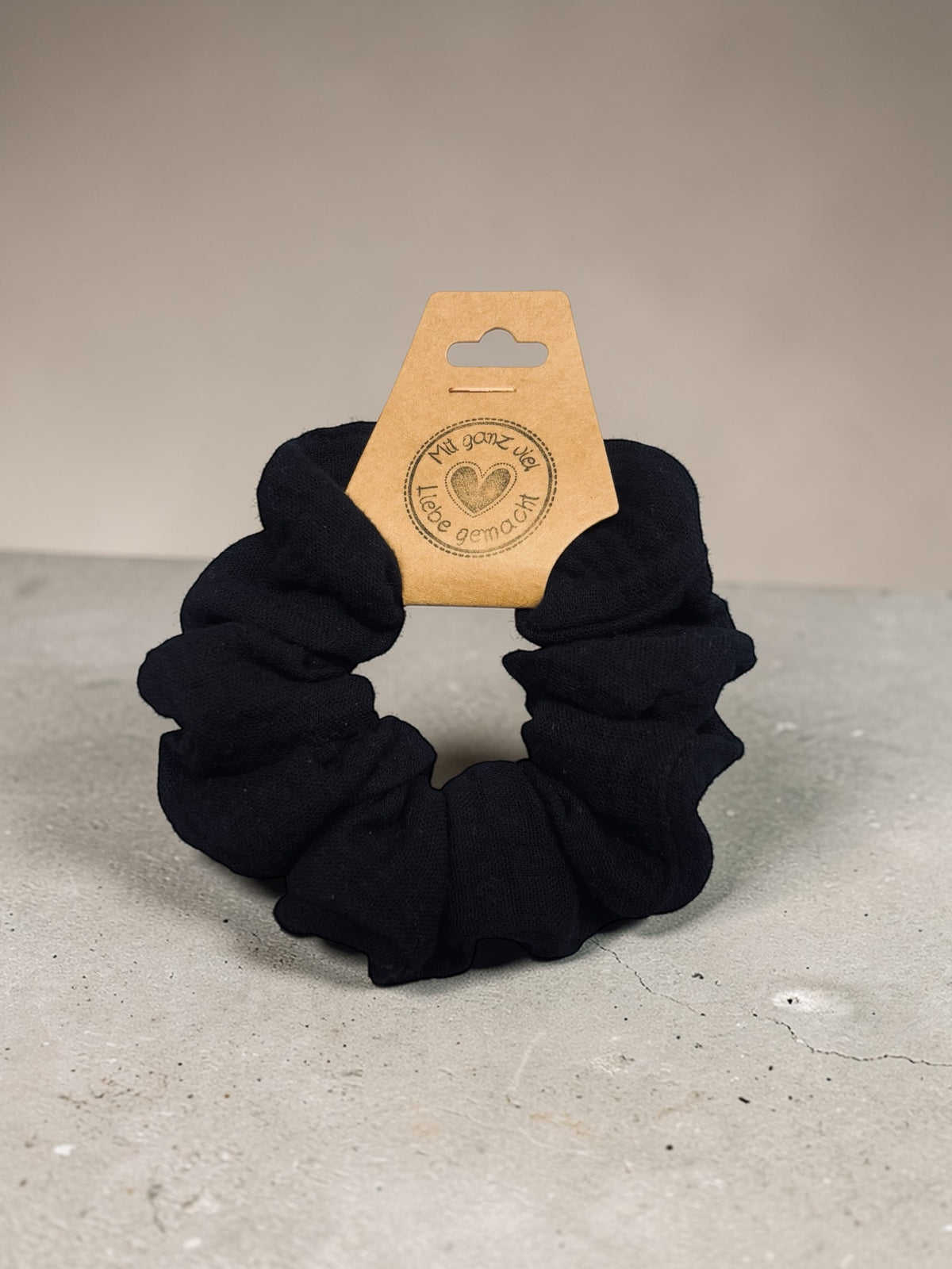 Großer handgemachter schwarzer Musselin-Scrunchie, ca. 10 cm Außendurchmesser, aufrecht mit Papieretikett auf hellem Untergrund
