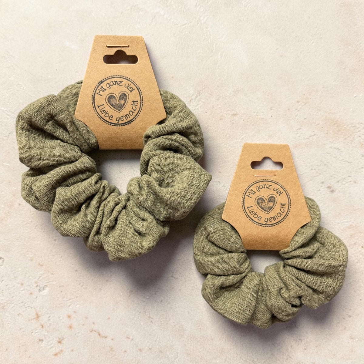 Zwei handgemachte Musselin-Scrunchies in Oliv, groß links und klein rechts, auf hellem Stein zum Größenvergleich
