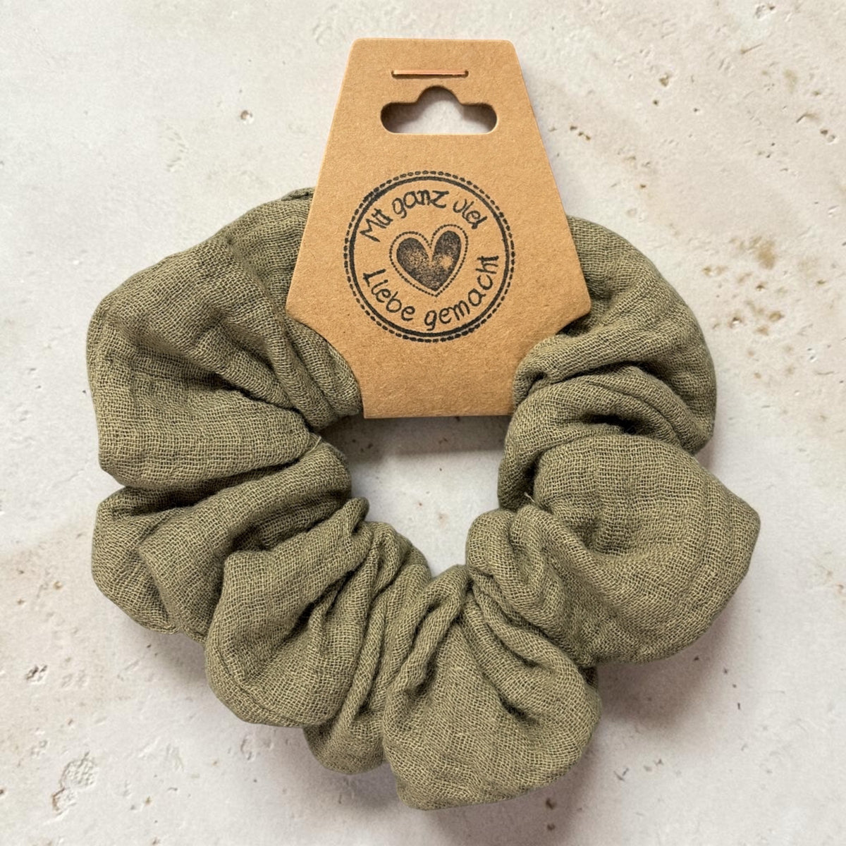 Großer handgemachter Musselin-Scrunchie in Oliv, ca. 10 cm Außendurchmesser, von oben mit Papieretikett auf hellem Stein
