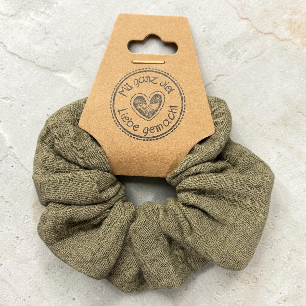 Kleiner handgemachter Musselin-Scrunchie in Oliv, ca. 8 cm Außendurchmesser, von oben mit Papieretikett auf hellem Stein
